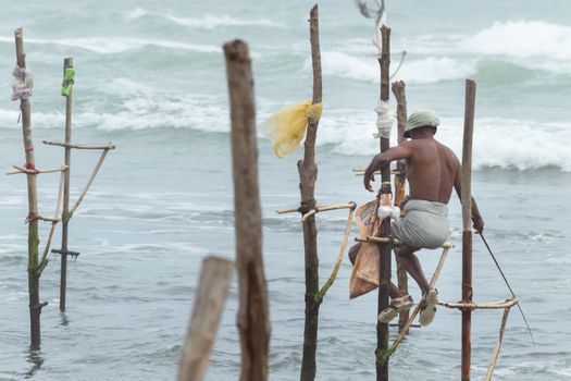 Old stilt fisherman with his wooden rod facing back to camera angel, fishing in a traditional unique method in Sri Lankan culture, sunny bright evening on the beach.