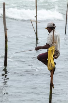 Stilt fisherman with his wooden rod facing side to the camera with his yellow pocket on the pole, fishing in a traditional unique method in Sri Lankan culture, sunny bright evening on the beach.