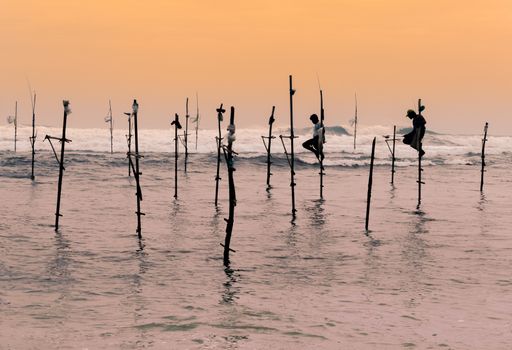 A couple of Stilt fishermen sitting on their poles with wooden fishing rods in their hands in the sunset evening. Ocean waves crash behind them in the background with an orange yellowish sky.