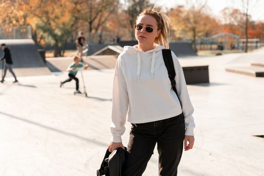 Outdoor portrait of young beautiful woman with a ponytail and sunglasses, with a backpack on his shoulders, dressed in a white sweater, near the sportsground. White hoodie.