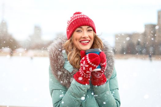 Beautiful lovely middle-aged girl curly hair warm winter jackets red knitted hat glove stands ice rink background Town Square Christmas mood lifestyle Happy holiday woman drink hot beverage coffee