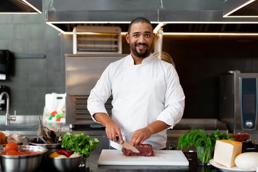 Handsome young African chef standing in professional kitchen in restaurant preparing a meal of meat and cheese vegetables. Portrait of man in cook uniform Cuts meat with a metal knife.