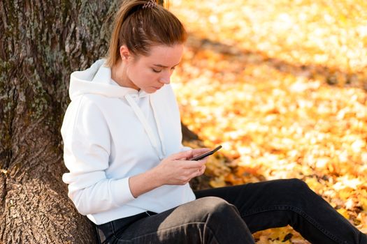 Woman in a white hoodie sweater with a hood sits on ground in the park and holding a cellphone in her hands. cute girl use smartphone for communication Female texting a message Communicaion concept