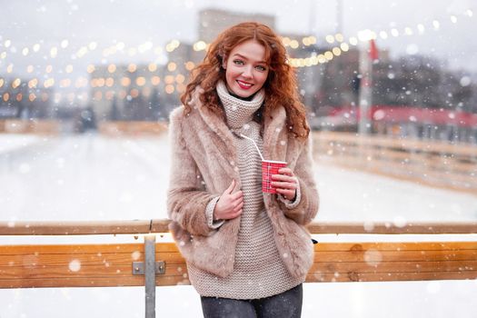 Young beautiful redhead girl freckles ice rink background Pretty woman curly hair portrait walking new year fair Modern cute female drink hot beverage mulled wine chrismas holiday decorated street.