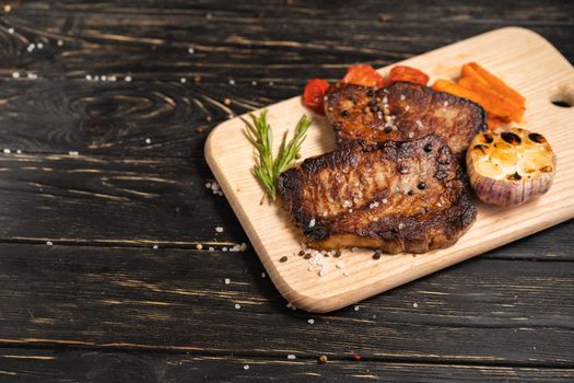 A juicy piece of fried meat with grilled cherry tomatoes garlic and carrot lies on a cutting board against a black wooden table. Degree of roasting well done. Food concept with copy space