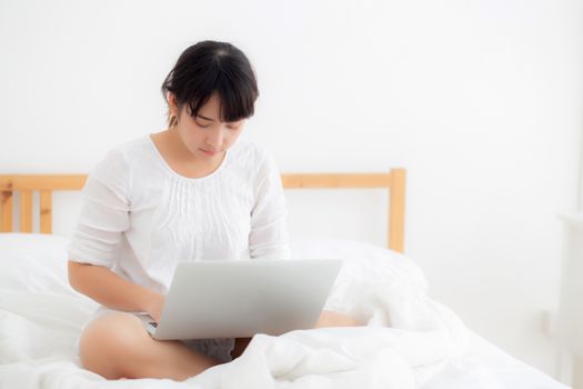 Portrait of beautiful asian young woman sitting on bed using laptop computer at bedroom for leisure and relax, freelance with girl working notebook, communication and lifestyle concept.