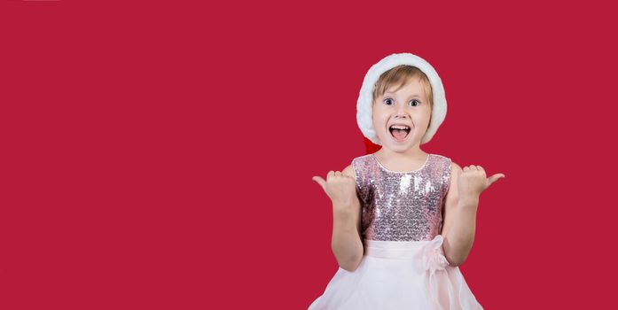 Surprised funny cacusian cute child girl in santa hat looking at the camera, shouting happily and showing thumbs up isolated on red background. Merry Christmas presents shopping sale..