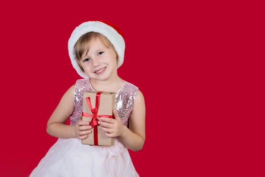 Surprised funny cacusian cute child girl in santa hat looking at the camera, smiling widly happily and hugs a gift isolated on red background. Merry Christmas presents shopping sale.