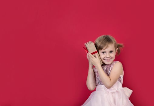 Smiling cute cacusian little girl in santa hat looking at the camera with gift box near her ear celebrating happy 2021 New Year isolated on red background. Merry Christmas presents shopping sale.