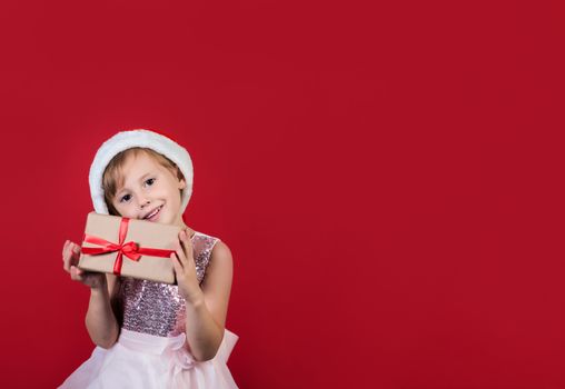 .Excited funny cacusian cute child girl in santa hat looking at camera holding gift box near face celebrating happy 2021 New Year isolated on red background. Merry Christmas presents shopping sale.