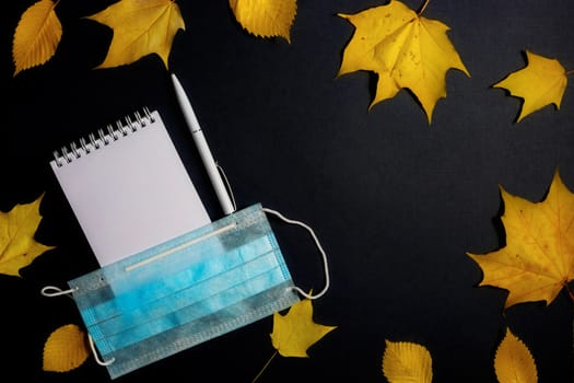 .Autumn fallen foliage, notebook and medical mask on black background.