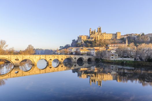 Under a winter blue sky in Béziers with its magnificent medieval bridge from the 12th century and its Gothic cathedral and their perfect reflection on the Orb river.