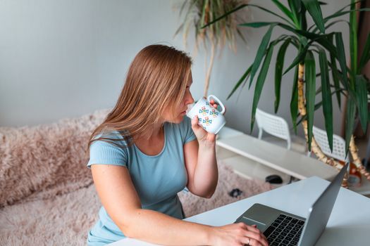 Middle age woman drinking tea sitting at the table at home working using computer laptop. Work from home and stay at home during coronovirus pandemic concept