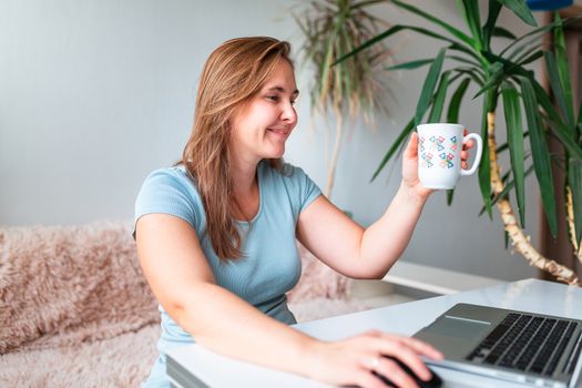 Middle age woman drinking tea sitting at the table at home working using computer laptop. Work from home and stay at home during coronovirus pandemic concept