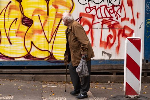 Old men is walking during the morning with his walking stick in Prague 7 while contemplating a graffiti during quarantine period due to outbreak of COVID-19 as winter is starting, Czech Republic
