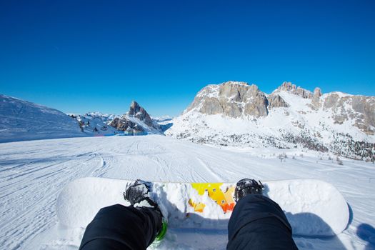 Snowboarder sitting on ski slope at resort in Dolomites Italian Alps view on legs and snowboard
