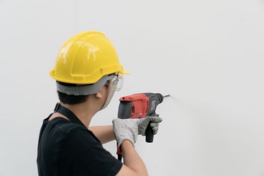 Man builder or worker with yellow helmet and goggle drilling machine on white wall.