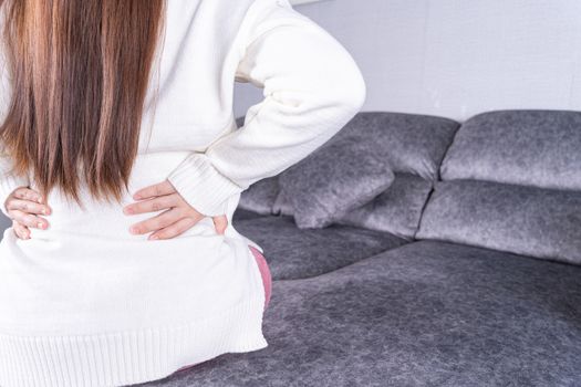 Young woman suffering from back pain while sitting on sofa at home. Healthcare medical or daily life concept.