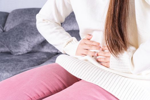 Young woman suffering from strong abdominal pain or menstruation while sitting on sofa at home. Healthcare medical or daily life concept.