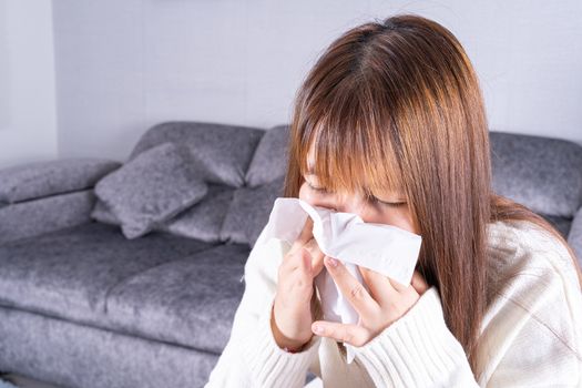 Asian beautiful woman blowing a nose to tissue while sitting on sofa at home. Healthcare medical or daily life concept.