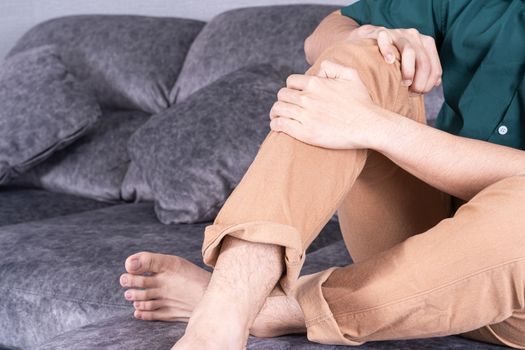 Young man suffering from knee pain while sitting on sofa at home. Healthcare medical or daily life concept.