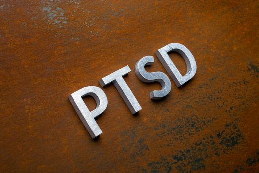 the word PTSD - post traumatic stress disorder - laid with silver metal letters on flat rusted sheet steel background in slanted diagonal perspective.