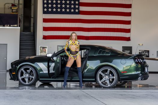 A gorgeous blonde model poses with an American muscle car inside an aircraft hangar