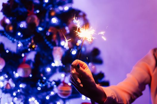female hand holds a sparkler on the background of a decorated Christmas tree. The concept of winter holidays and fun.