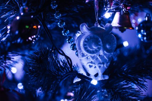 Decorated Christmas tree close-up in neon blue light. Toy glass owl and garland with lights with lanterns. New Year's baubles macro photo with bokeh. Winter holiday light decoration