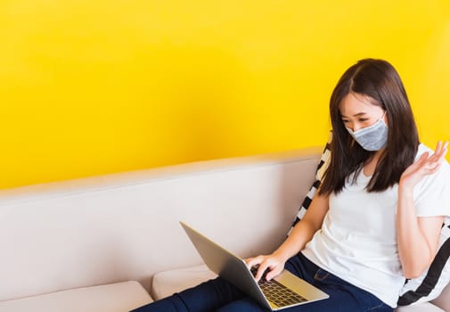 Portrait Asian of beautiful young woman sitting on sofa wearing medical face mask protective she work from home with laptop computer during Coronavirus studio shot isolated on yellow background