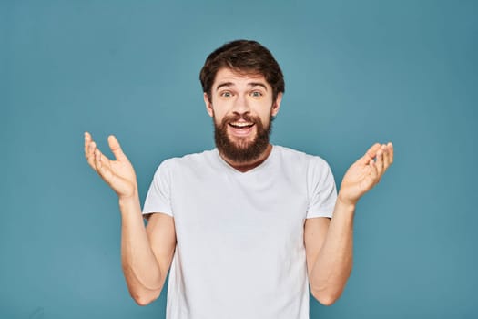 A bearded man in a white T-shirt gestures with his hands emotions blue background. High quality photo