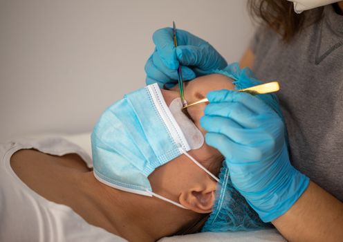 Treatment beauty procedure of upgrade Eyelashes Extension during a pandemic with preventive measures. Woman face wirh protective cap and facemask