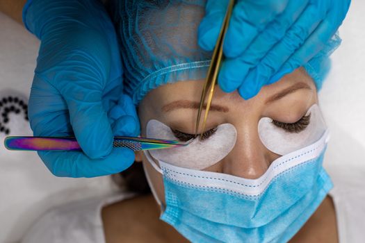 Treatment beauty procedure of upgrade Eyelashes Extension during a pandemic with preventive measures. Woman face wirh protective cap and facemask