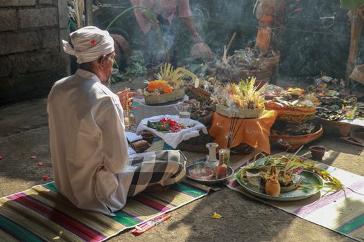 Rear view to Hindu priest praying during a wedding ceremony. Pedanda sits on the ground in front of the offerings and performs wedding ceremonies.