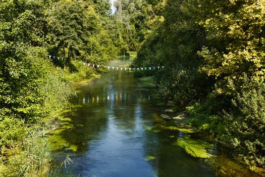 Scenic view of rural river with colored flags hanging with rope , trees and wild plants along the riverbanks