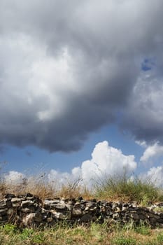 Italian rural landscape with low stone wall made of irregular broken stones , beautiful blue sky with clouds ,  green and dry grass in the foreground 