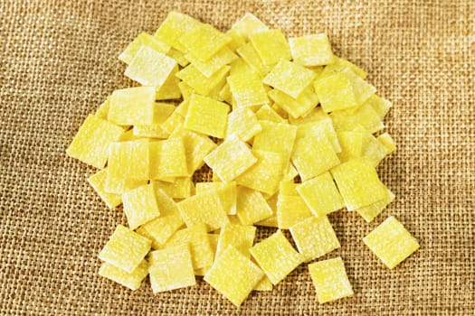 Pile of small flat squares of egg pasta called quadrucci on sackcloth background,yellow color pasta with rough surface 
