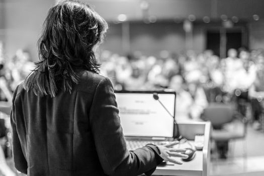 Female speaker giving a talk on corporate business conference. Unrecognizable people in audience at conference hall. Business and Entrepreneurship event.