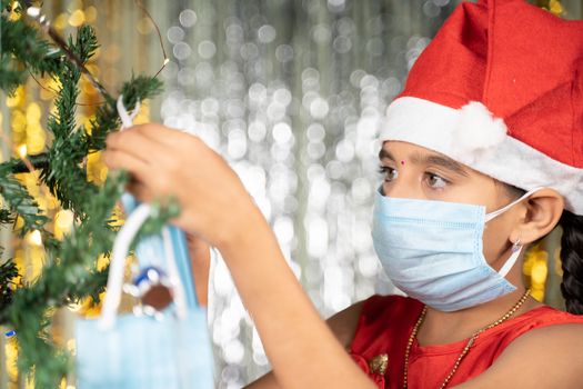 Girl kid in Medical mask and Santa Hat decorating Christmas tree - Concept of Covid-19, Coronavirus and Christmas Holiday celebration