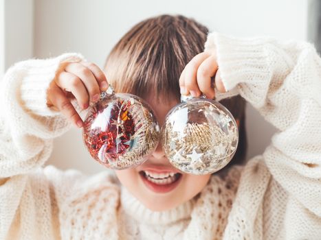 Kid with decorative balls for Christmas tree.Boy in cable-knit oversized sweater.Cozy outfit for snuggle weather.Transparent balls with red, golden spangles inside.Winter holiday spirit.New year.