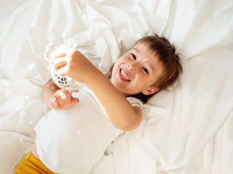 Little boy plays with sparkling mirror disco ball in bed and laughing happily. Joyful toddler. Playful child and sunny morning in cozy home.