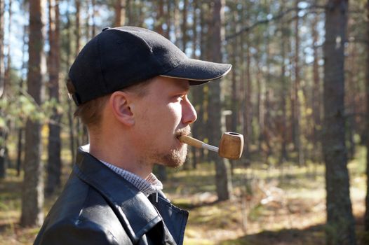 young man in a cap smokes a pipe in the forest, portrait