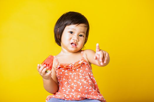 Happy portrait Asian baby or kid cute little girl attractive laugh smile wearing t-shirt playing holds cut watermelon fresh for eating, studio shot isolated on yellow background, healthy food and summer concept