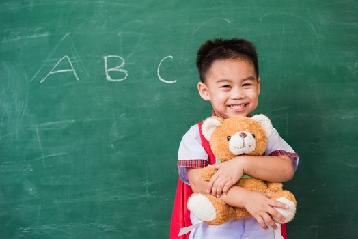 Back to School. Happy Asian funny cute little child boy from kindergarten in student uniform with school bag smiling and hugging teddy bear on green school blackboard, First time to school education