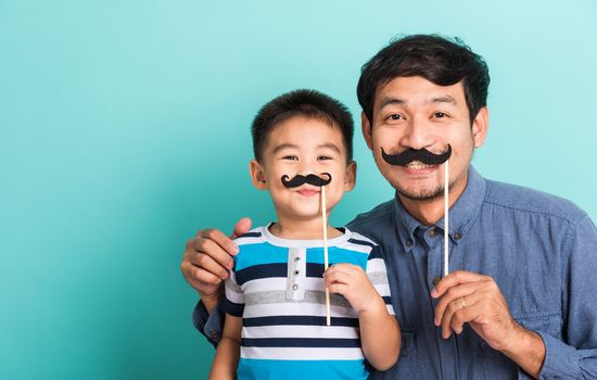 Family funny happy hipster father and his son kid holding black mustache props for the photo booth close face, studio shot isolated on a blue background, November men health awareness