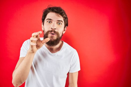 A man in a white T-shirt with a beard gestures with his hands emotions red background. High quality photo
