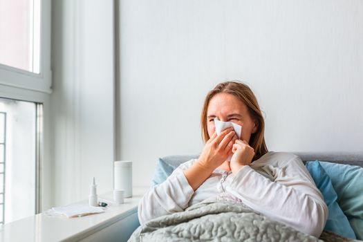 Sick woman blowing her nose with headache and fever lying under the blanket. Sick woman staying in bed with temperature durong coronavirus pandemic. Sick woman covered with a blanket lying in bed with high fever and a flu, resting.