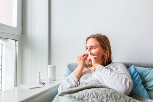 Sick woman blowing her nose with headache and fever lying under the blanket. Sick woman staying in bed with temperature durong coronavirus pandemic. Sick woman covered with a blanket lying in bed with high fever and a flu, resting.