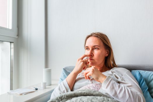 Sick woman spraying her nose with headache and fever lying under the blanket. Sick woman staying in bed with temperature durong coronavirus pandemic. Sick woman covered with a blanket lying in bed with high fever and a flu, resting.