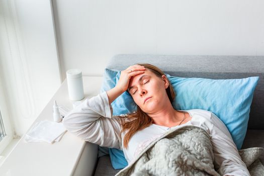 Sick woman with headache and fever lying under the blanket. Sick woman staying in bed with temperature durong coronavirus pandemic. Sick woman covered with a blanket lying in bed with high fever and a flu, resting.